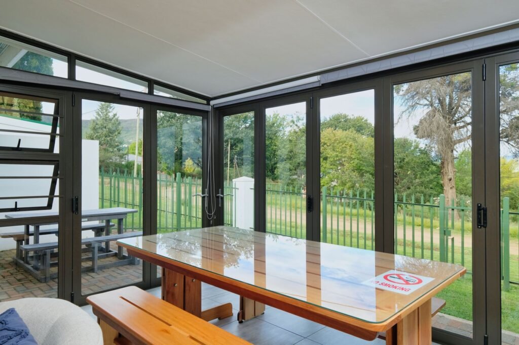Sistema de cerramiento de terraza con paneles de cristal y perfiles de aluminio oscuro abiertos hacia el jardín, mostrando el sistema abatible en posición abierta.