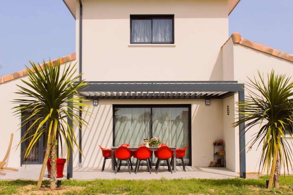Pérgola bioclimática adosada a fachada de chalet con comedor exterior y sillas rojas