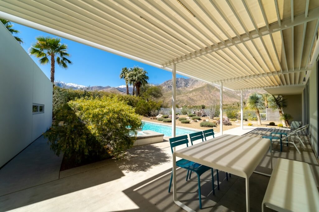 Pérgola bioclimática adosada a fachada de vivienda con lamas orientables, mesa de comedor exterior y piscina al fondo